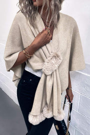 Taupe knit cape with fur trim and faux fur pom-poms, draped open over a white top.