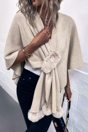 Taupe knit cape with fur trim and faux fur pom-poms, draped open over a white top.