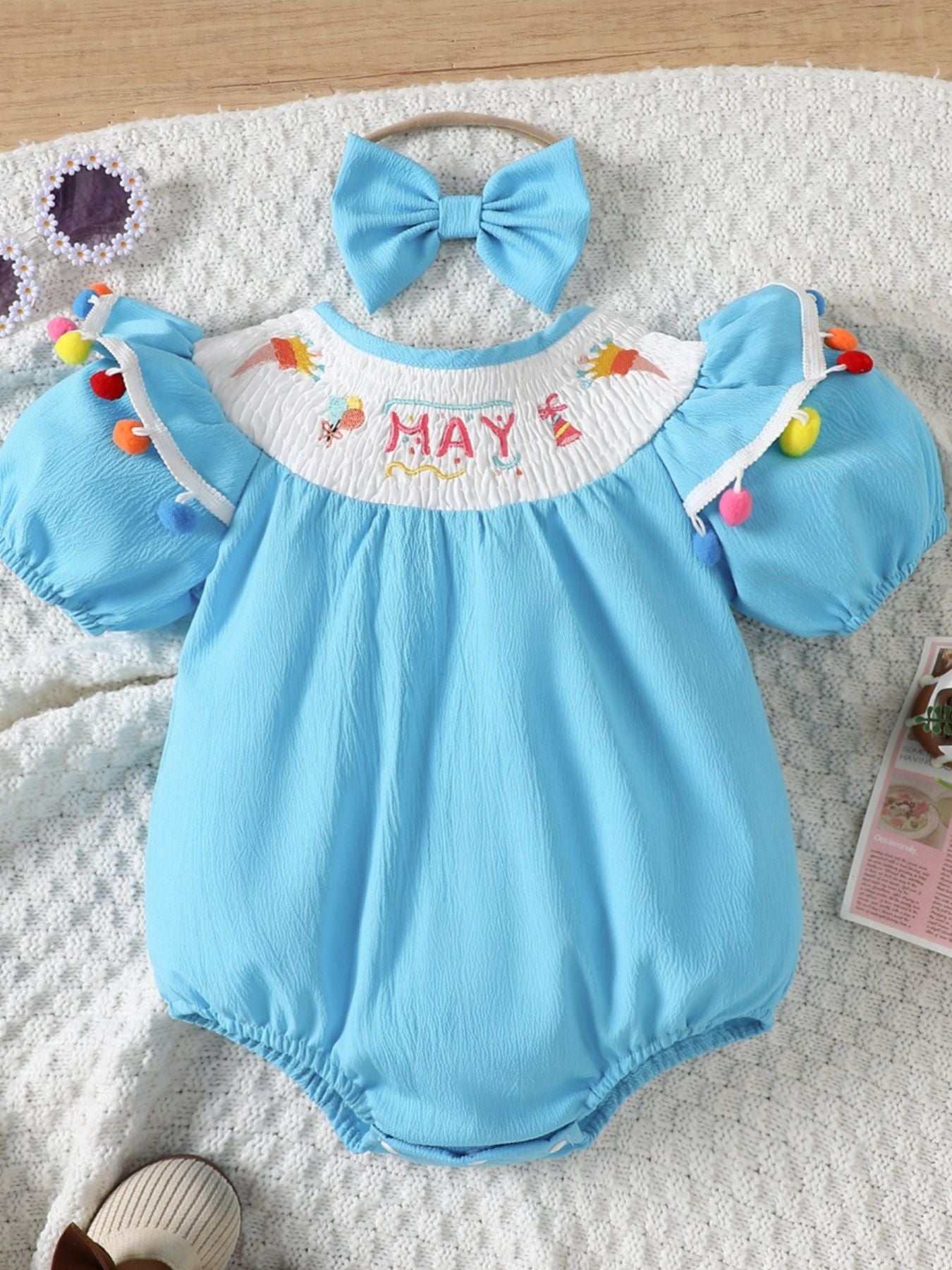 Pastel blue cotton baby romper with smocked neckline, "MAY" embroidery, puff sleeves, and pom-pom trim.