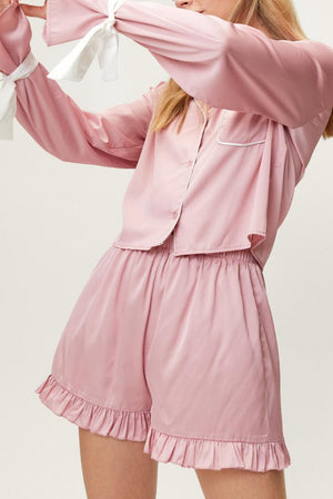 Blush pink silk pajama set with white piping, tie-sleeve detail, and ruffled shorts.