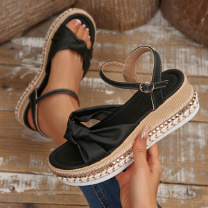 Black bow knot wedge sandals with ankle buckle and braided trim detail.