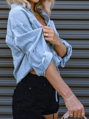 Light wash denim blouson shirt with rolled sleeves and buttoned front, styled with black shorts.