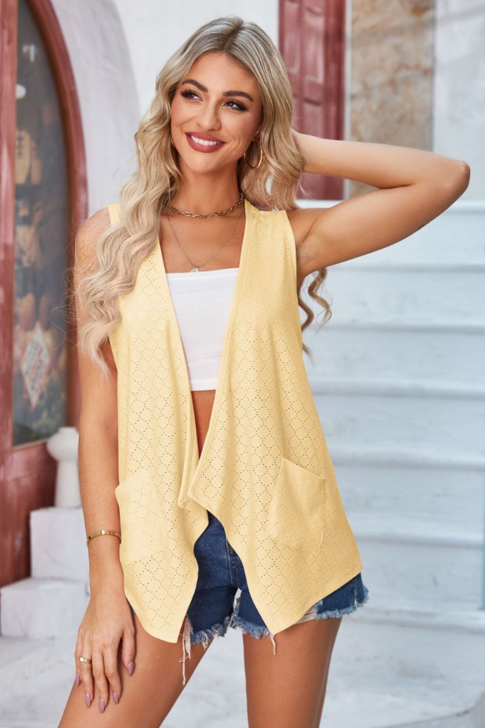 Yellow eyelet drape front vest with pockets and open front, styled with a white top and denim shorts.