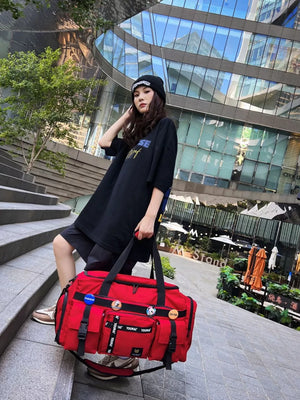 Red duffel bag with multiple pockets and decorative pins, held by a person wearing a black beanie and oversized t-shirt.