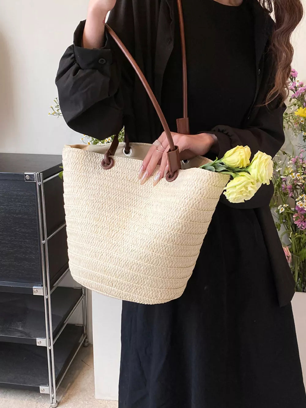 Natural straw woven large shoulder bag with brown leather straps, holding yellow flowers.
