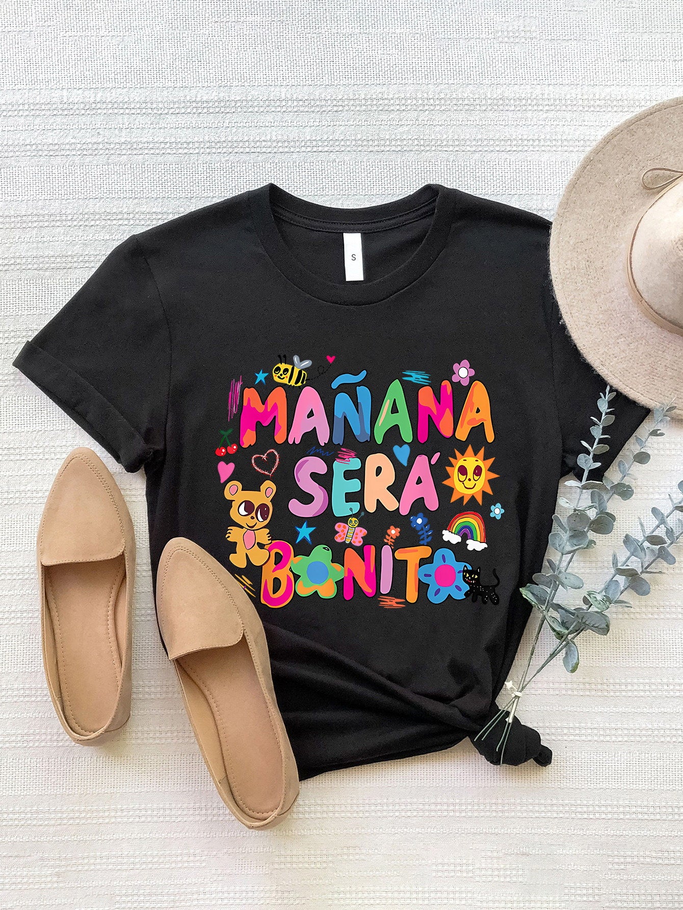 Black graphic tee with colorful "Mañana Sera Bonito" text, bee, teddy bear, sun, rainbow, and flowers.