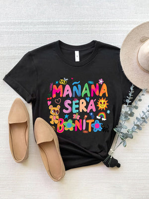 Black graphic tee with colorful "Mañana Sera Bonito" text, bee, teddy bear, sun, rainbow, and flowers.