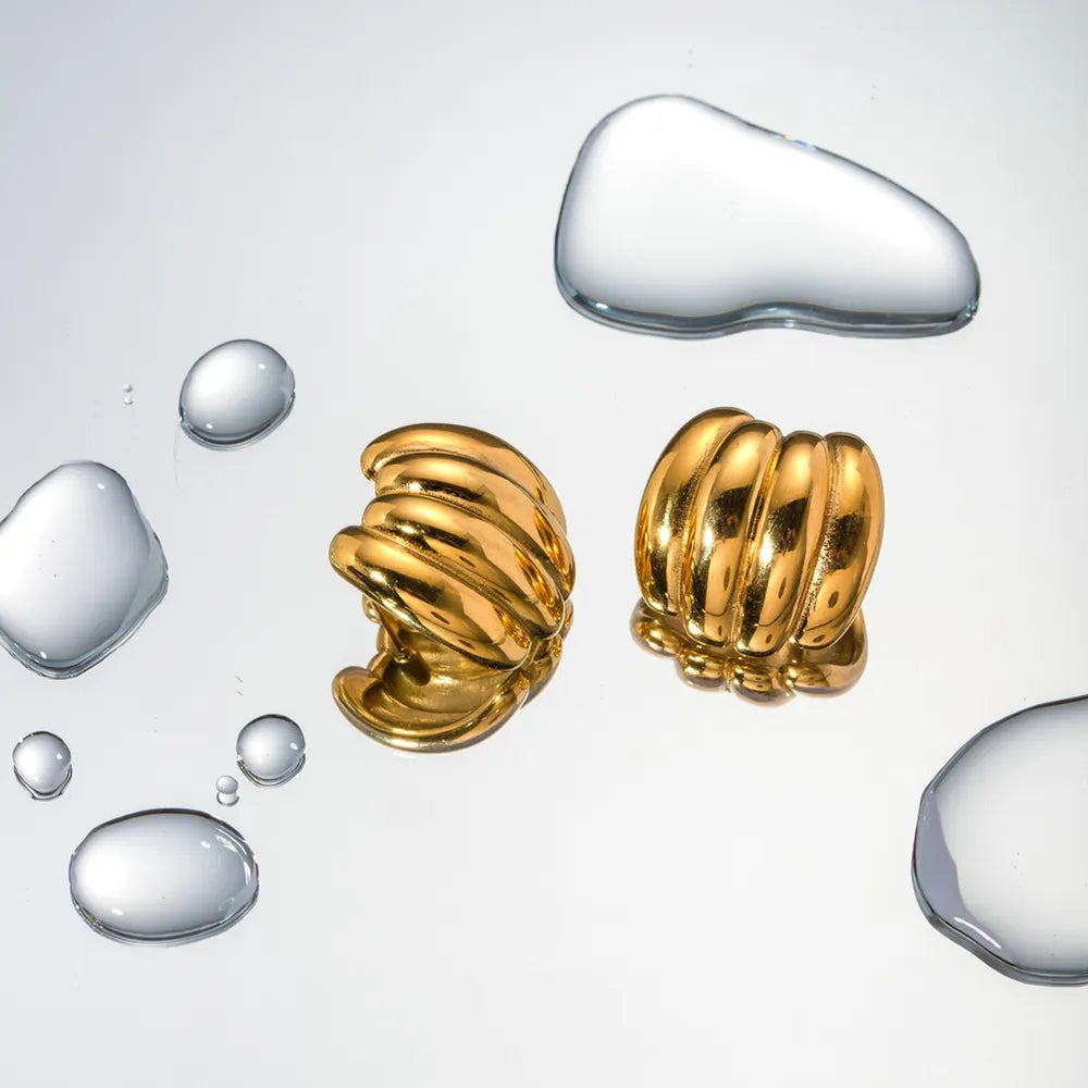 Sculptural ribbed gold earrings with 18K gold plating, displayed with water droplets.