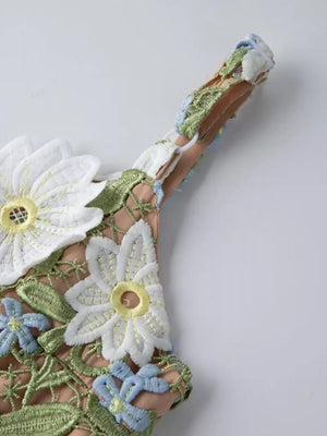 Embroidered floral maxi dress detail with white and blue flowers and green leaves on a tan background.
