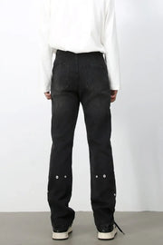 Dark wash denim trousers with asymmetric seams and decorative grommets on the lower legs.