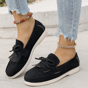 Black suede loafer flats with fringe and bow detail, styled with distressed jeans.