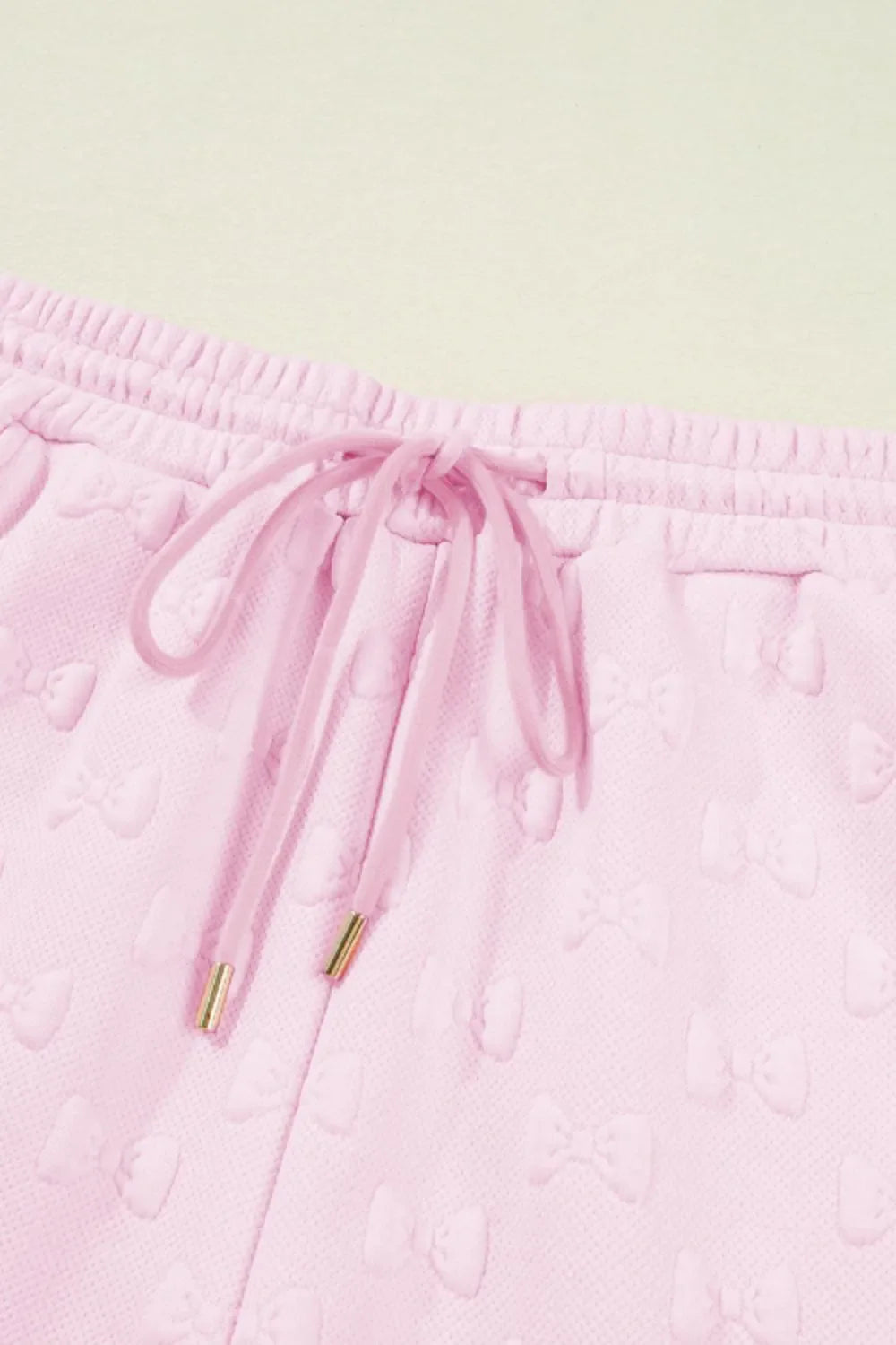 Blush pink lounge shorts with embossed bow pattern, drawstring waist, and gold-tipped aglets.
