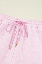 Blush pink lounge shorts with embossed bow pattern, drawstring waist, and gold-tipped aglets.