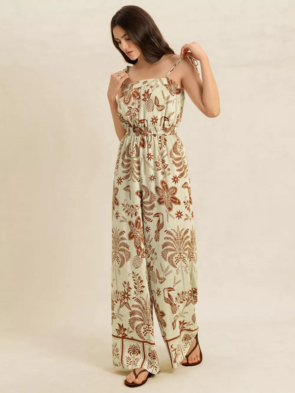 Tropical tie strap wide-leg romper in light green with brown floral and toucan print, smocked waist, and tie straps.