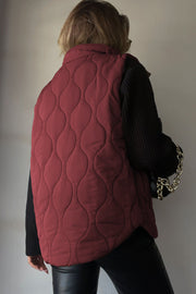 Wine quilted stand-collar gilet with wave pattern, worn over black sweater and pants.