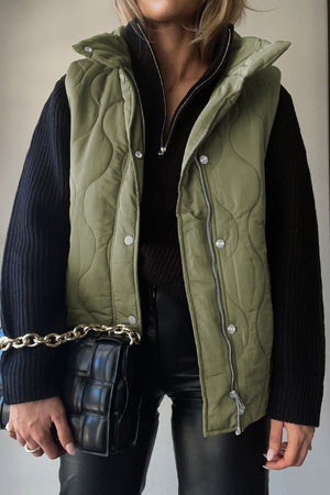 Green quilted stand-collar utility gilet with snap and zip closure, worn over a black sweater.