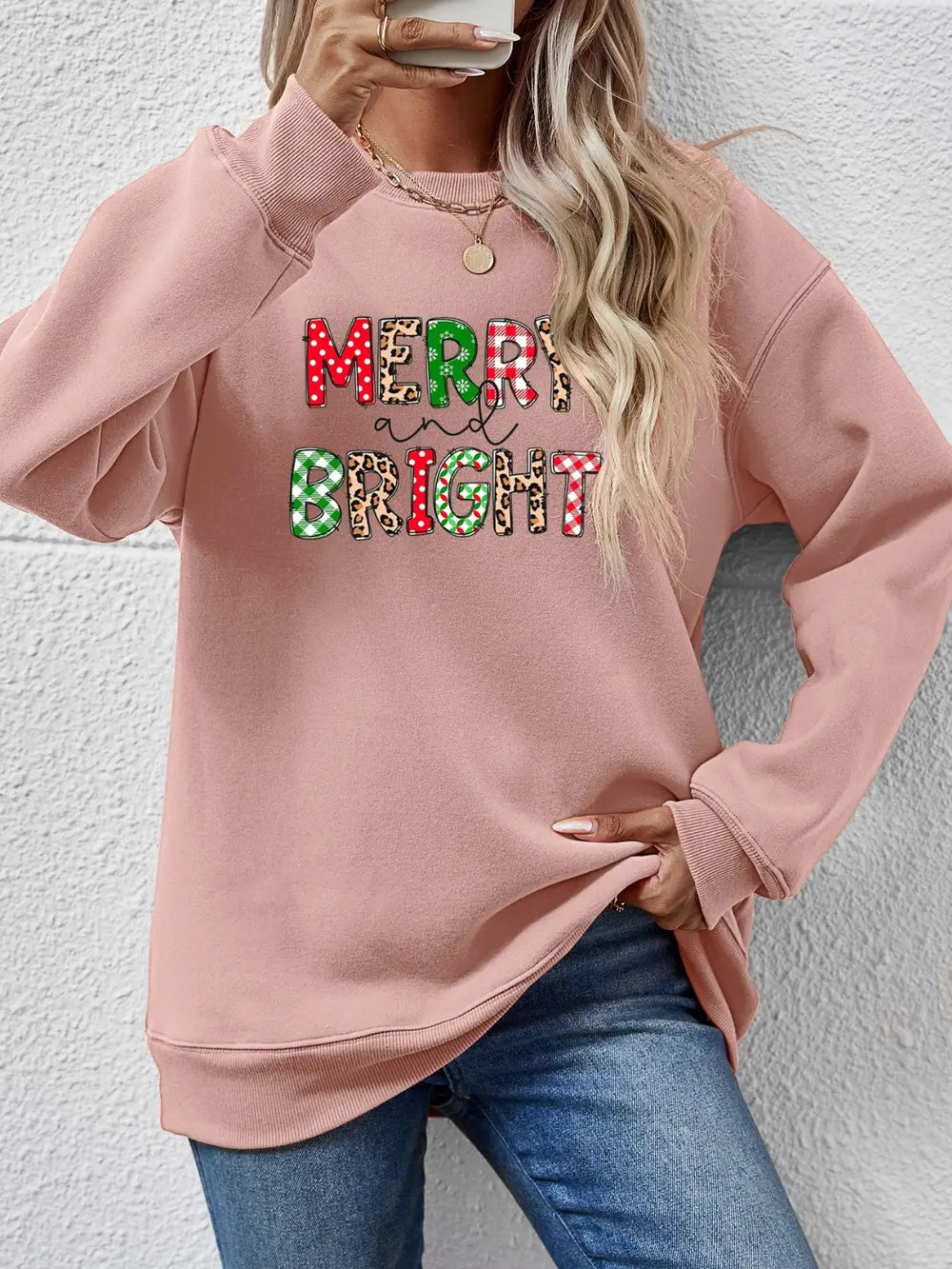 Blush pink crewneck sweatshirt with "Merry and Bright" graphic in festive patterns.