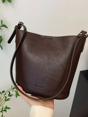 Distressed vintage bucket crossbody bag in brown with adjustable strap and embossed logo.