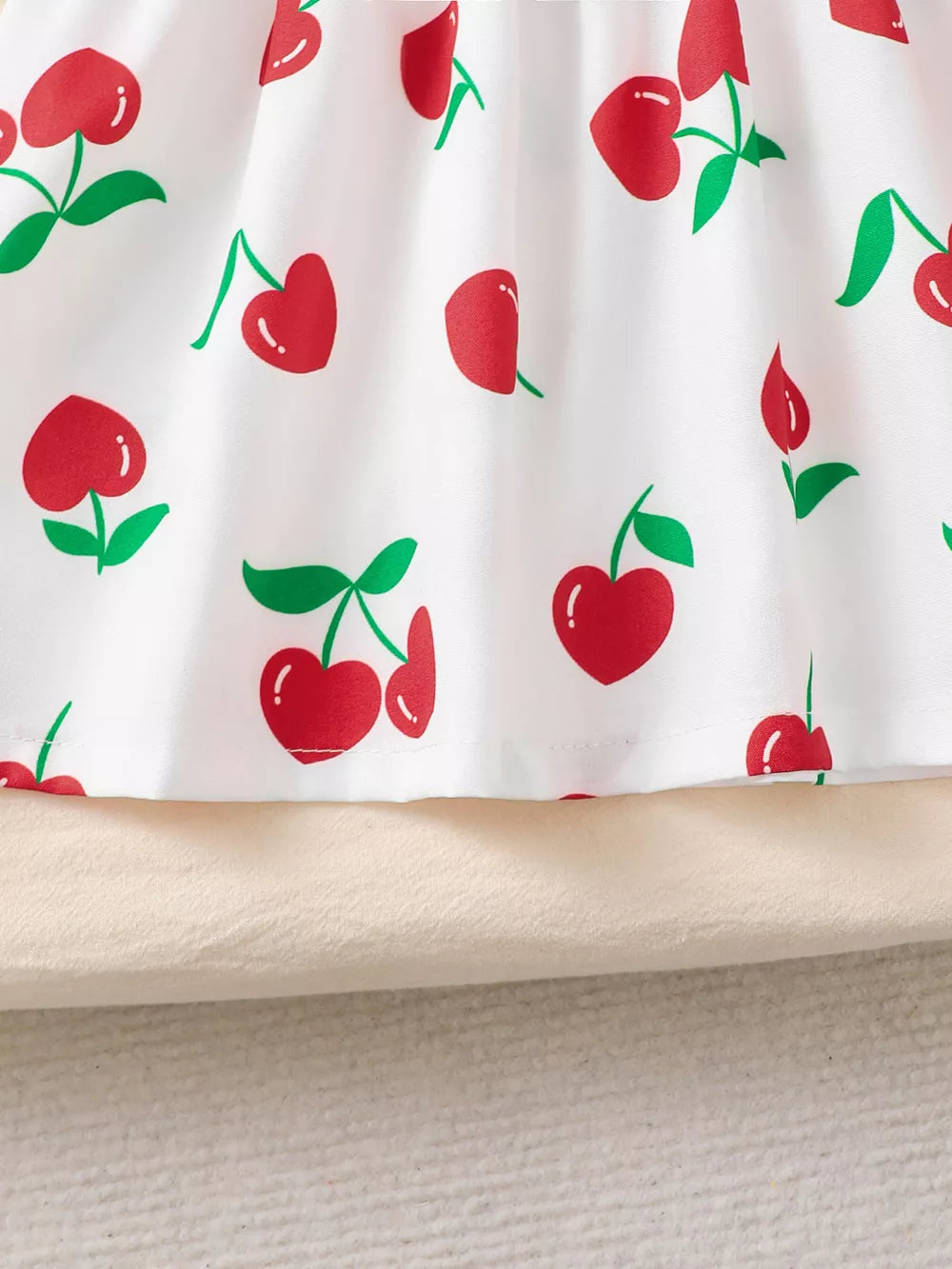Baby dress with white fabric and red cherry print, ruffled hem detail.