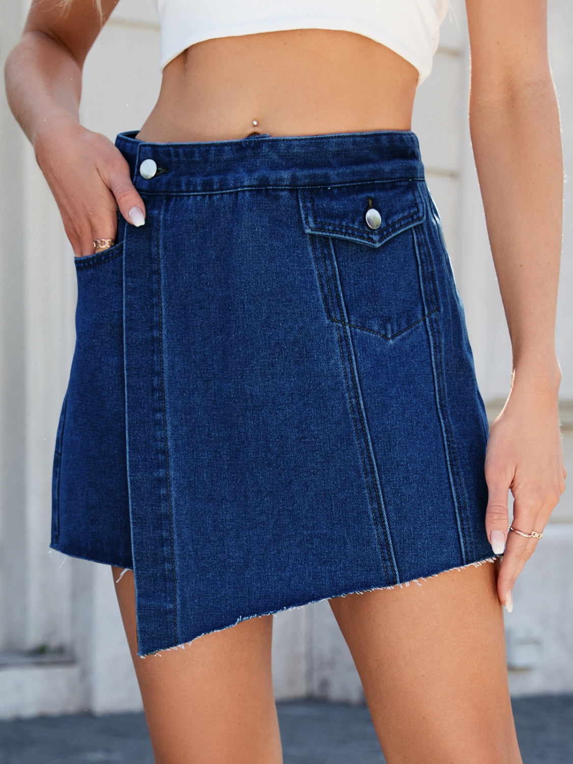 Dark wash denim skort with asymmetric wrap, raw hem, and pocket details.