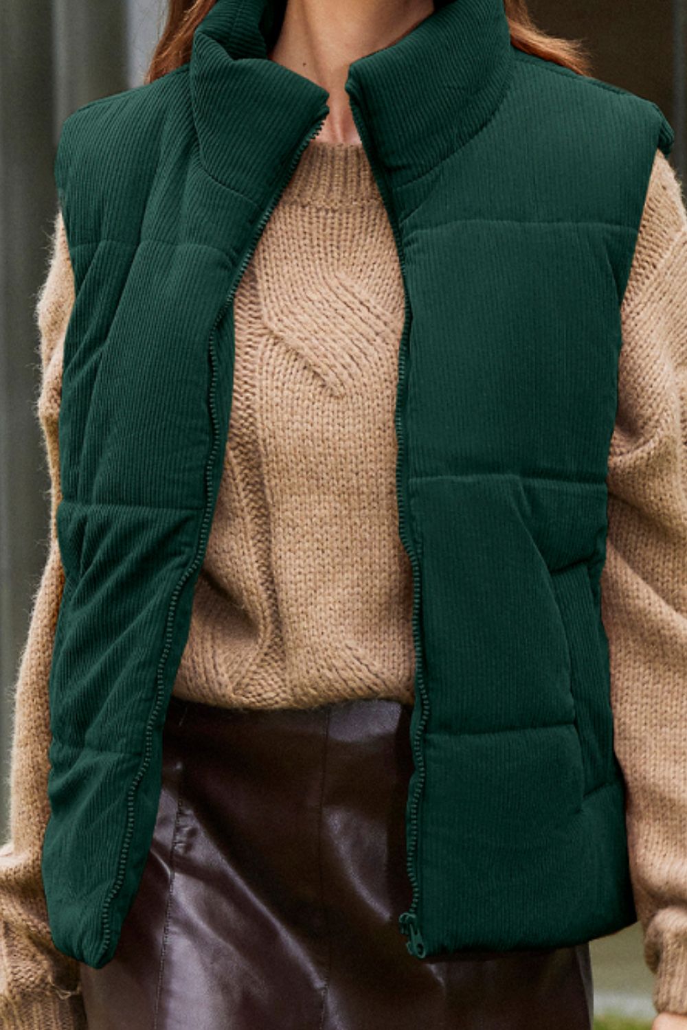 Dark green corduroy padded vest with a high collar and zipper closure, styled over a tan sweater.