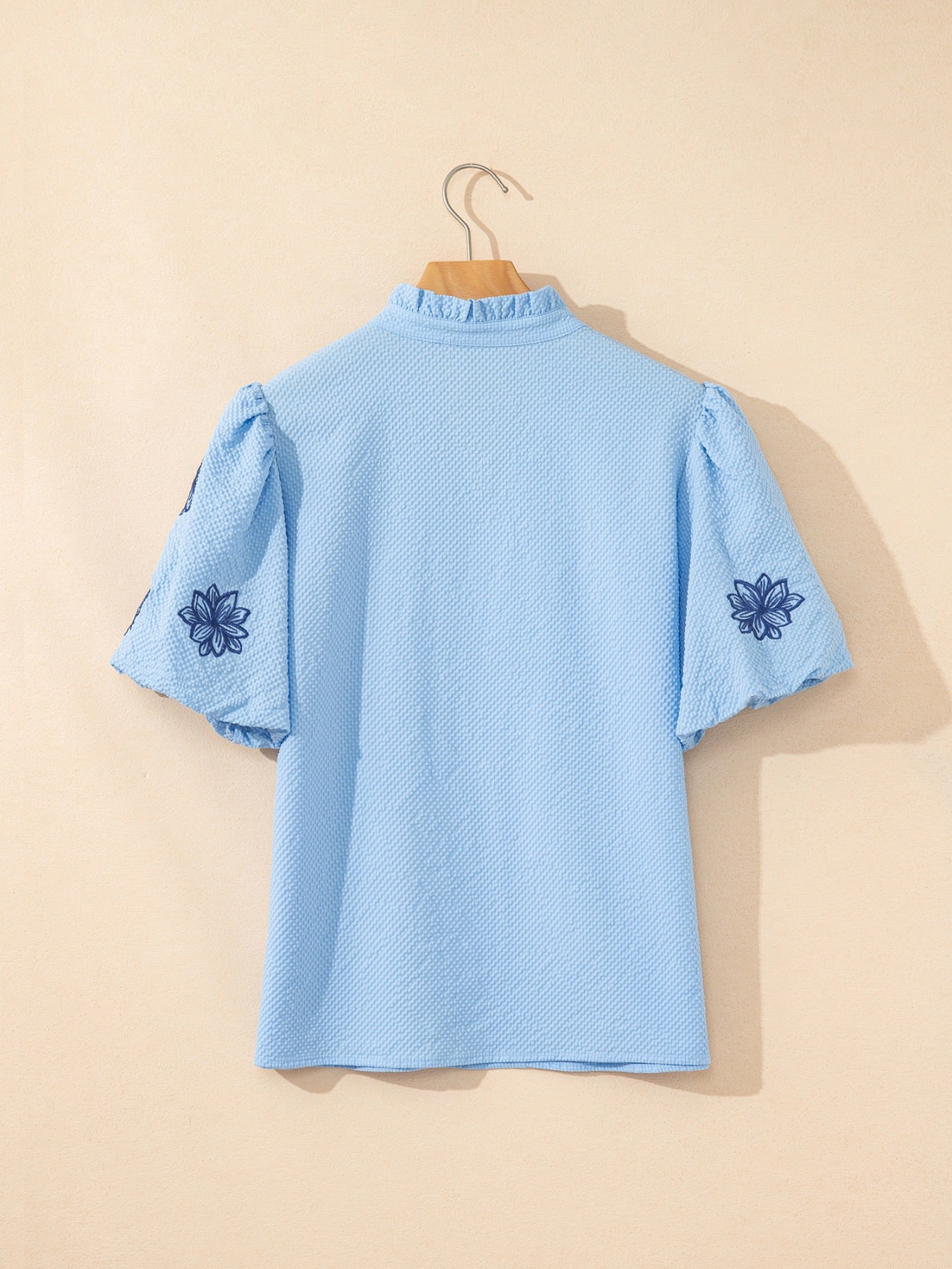 Light blue textured blouse with embroidered floral detail on puff sleeves and ruffled neckline.