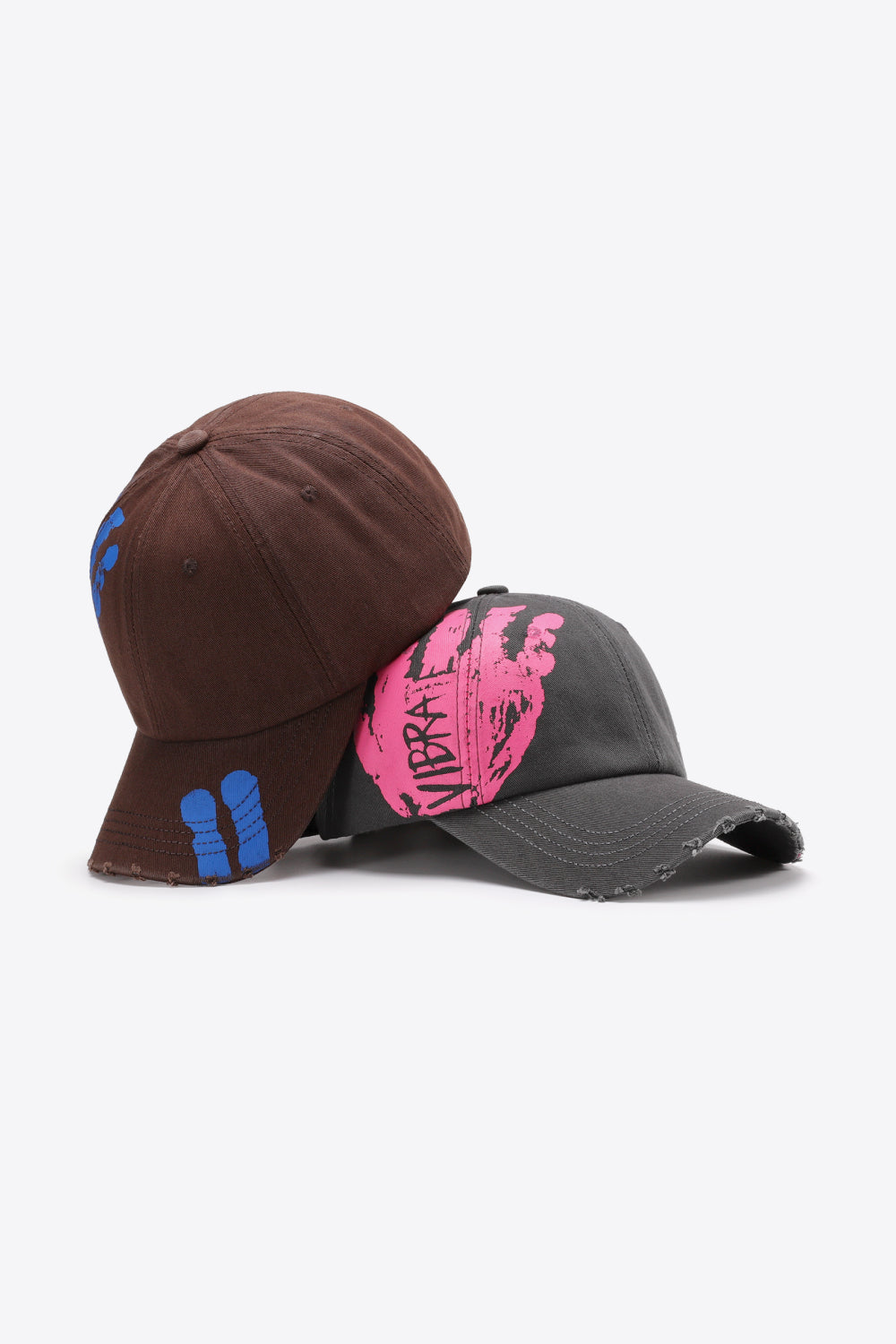 Dark gray baseball cap with pink graffiti art and distressed brim, and a brown cap with blue graffiti accents.