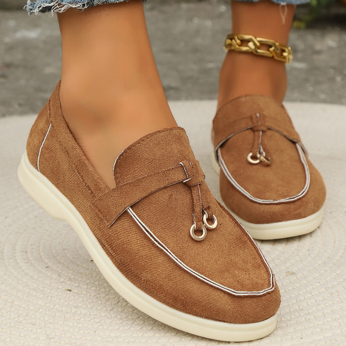 Brown flat slip-ons with decorative strap and tassel detail.