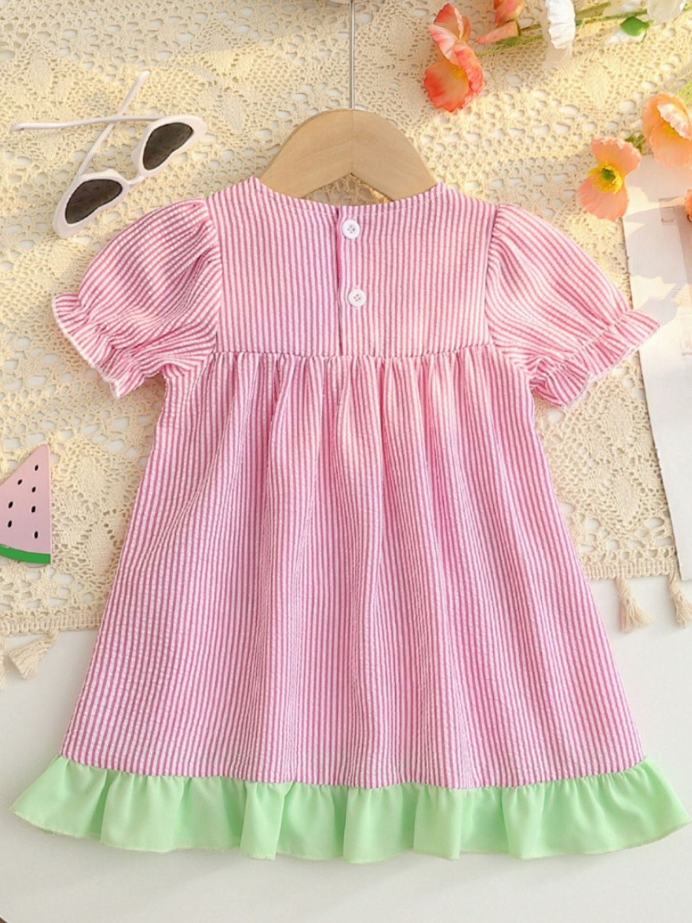 Baby dress in blush pink seersucker with white stripes, ruffled sleeves, and green hem.