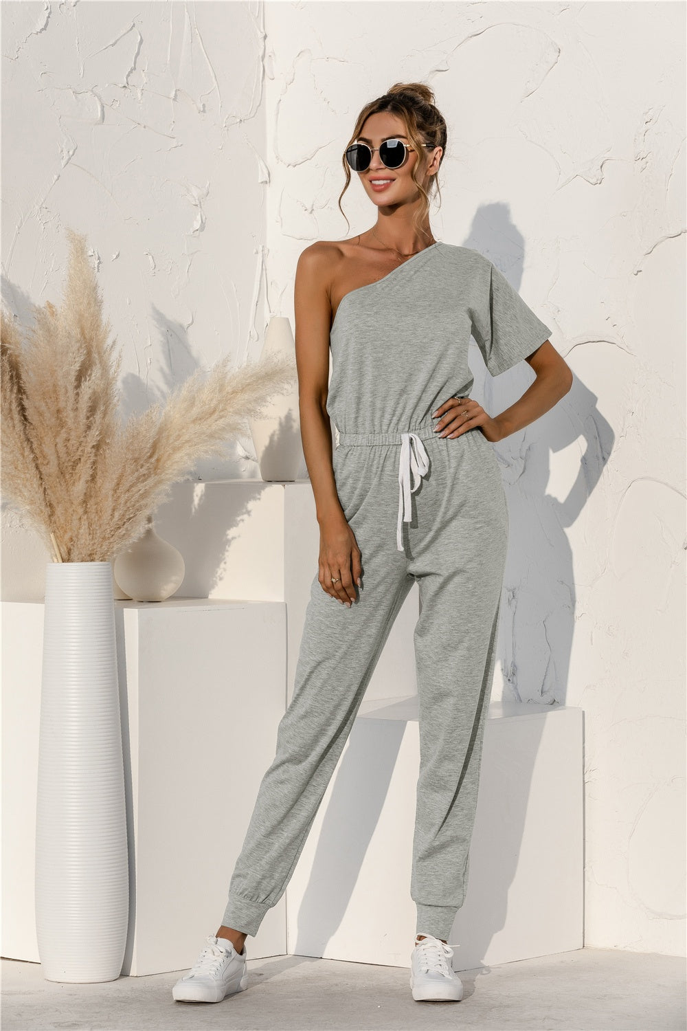 Gray asymmetric sweat jumpsuit with drawstring waist and cuffed jogger legs.
