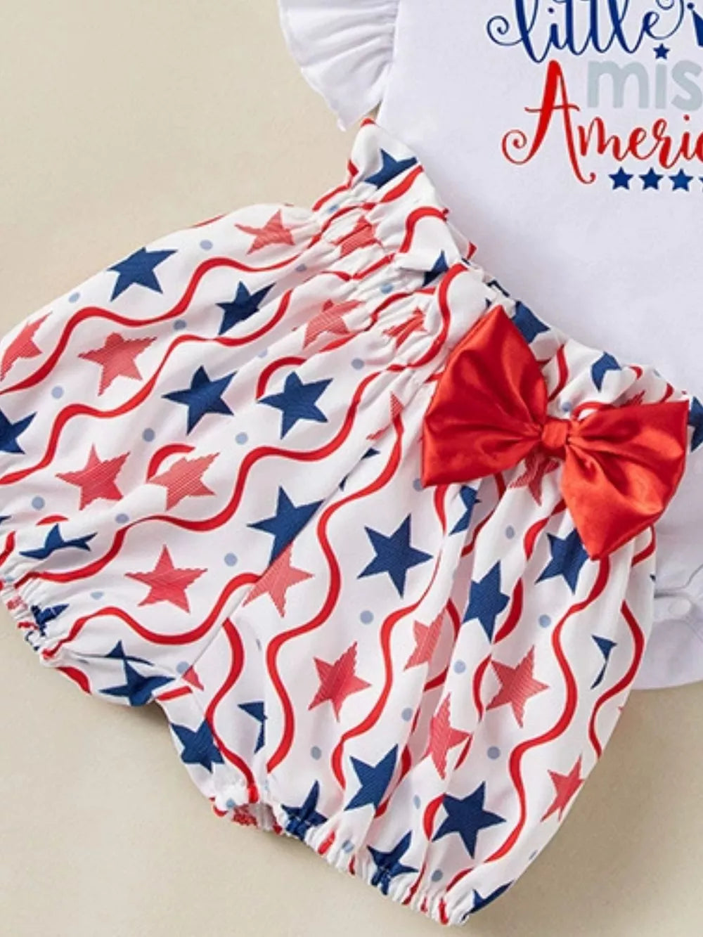 Baby girl outfit with red, white, and blue stars and wavy lines pattern, featuring bloomer shorts and a red bow.