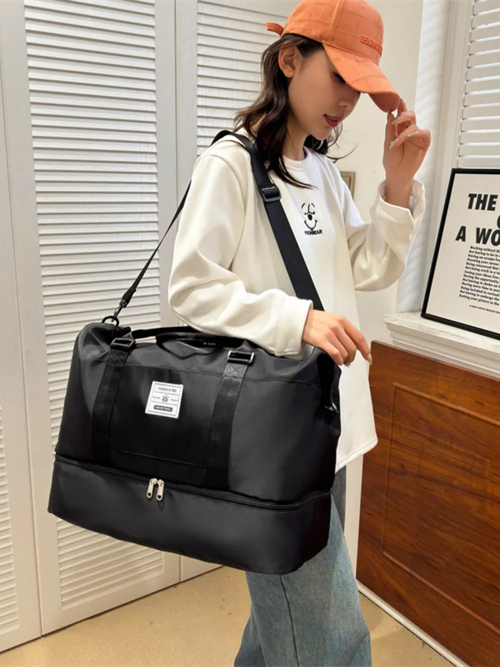 Black duffel bag with two compartments and a white label, worn crossbody by a woman.