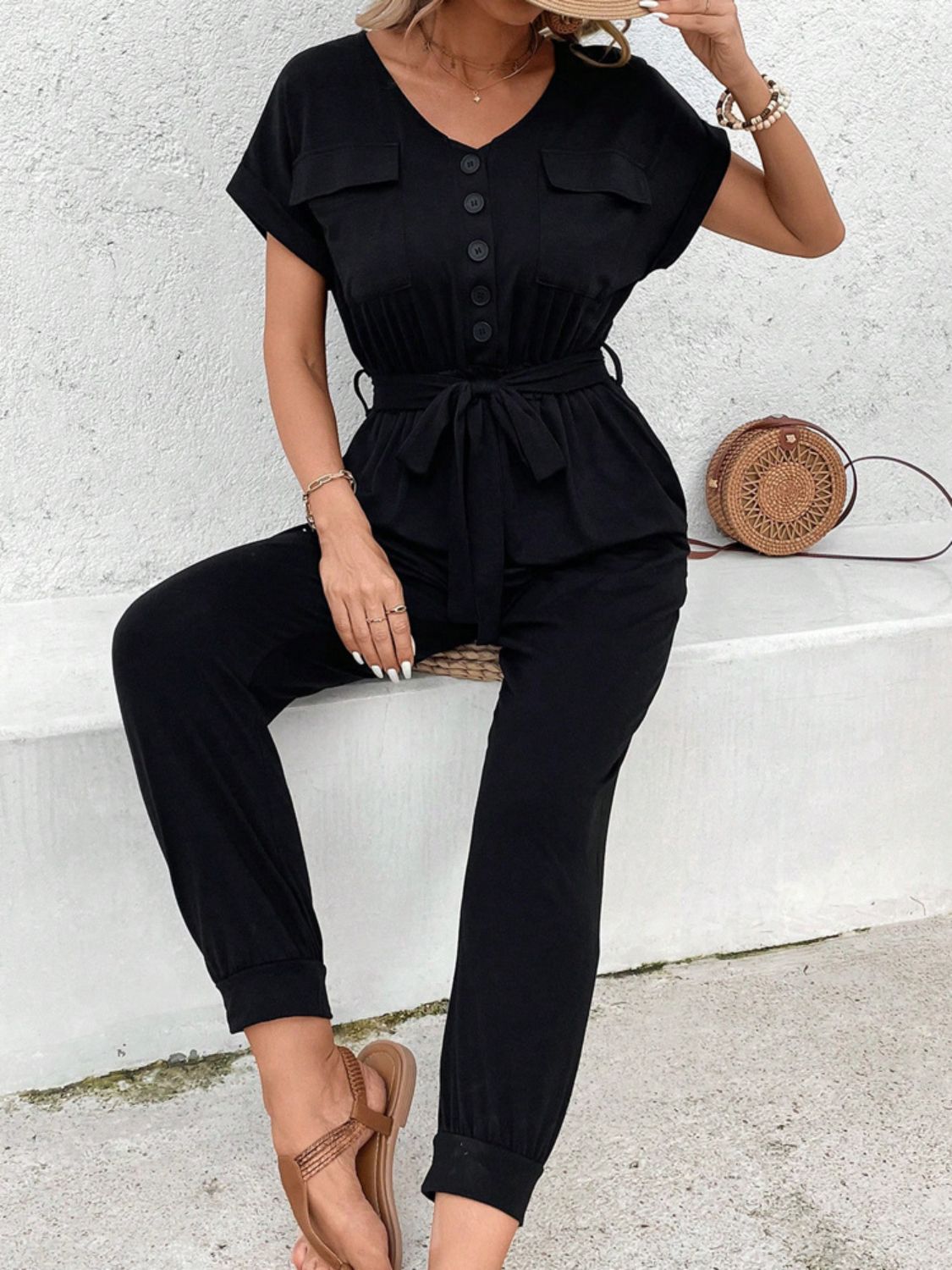 Black utility jumpsuit with tie waist, button front, V-neck, short sleeves, and chest pockets.