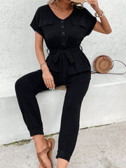 Black utility jumpsuit with tie waist, button front, V-neck, short sleeves, and chest pockets.