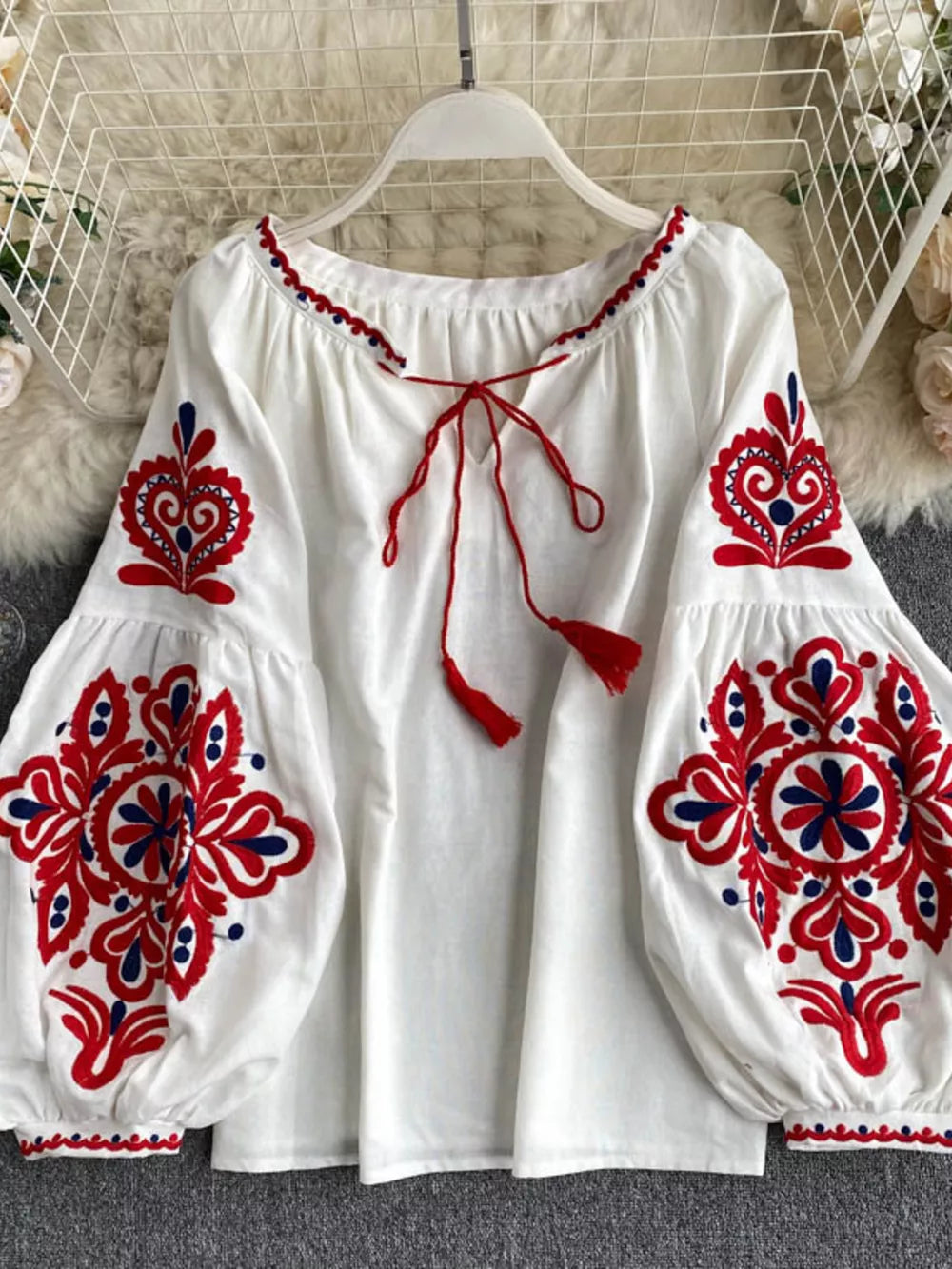 White peasant blouse with red and blue embroidery, tie neck, and tassel details.