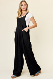 Black pleated bib wide leg jumpsuit with pockets, styled with a white tee.