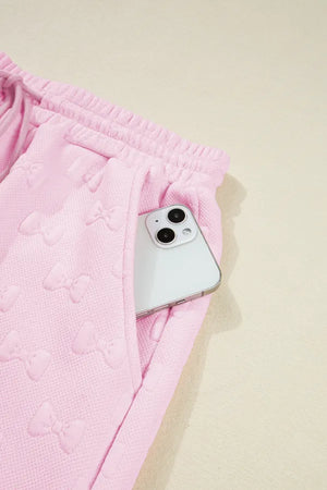 Blush pink loungewear set with embossed bow pattern and side pocket holding a phone.