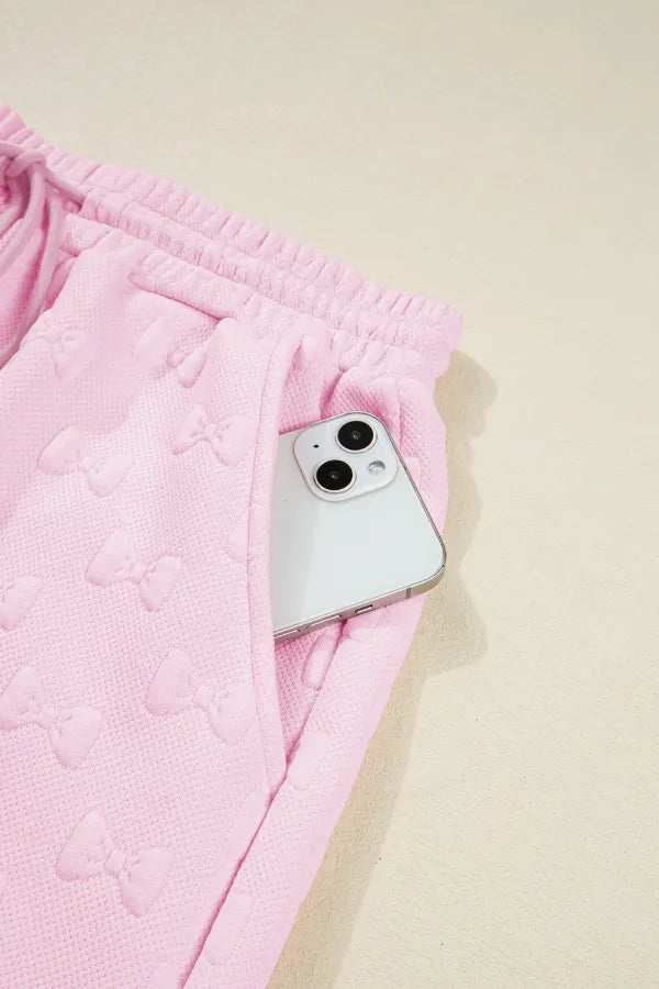 Blush pink loungewear set with embossed bow pattern and side pocket holding a phone.