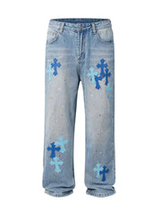 Light wash denim jeans with embroidered cross patches and paint splatters.
