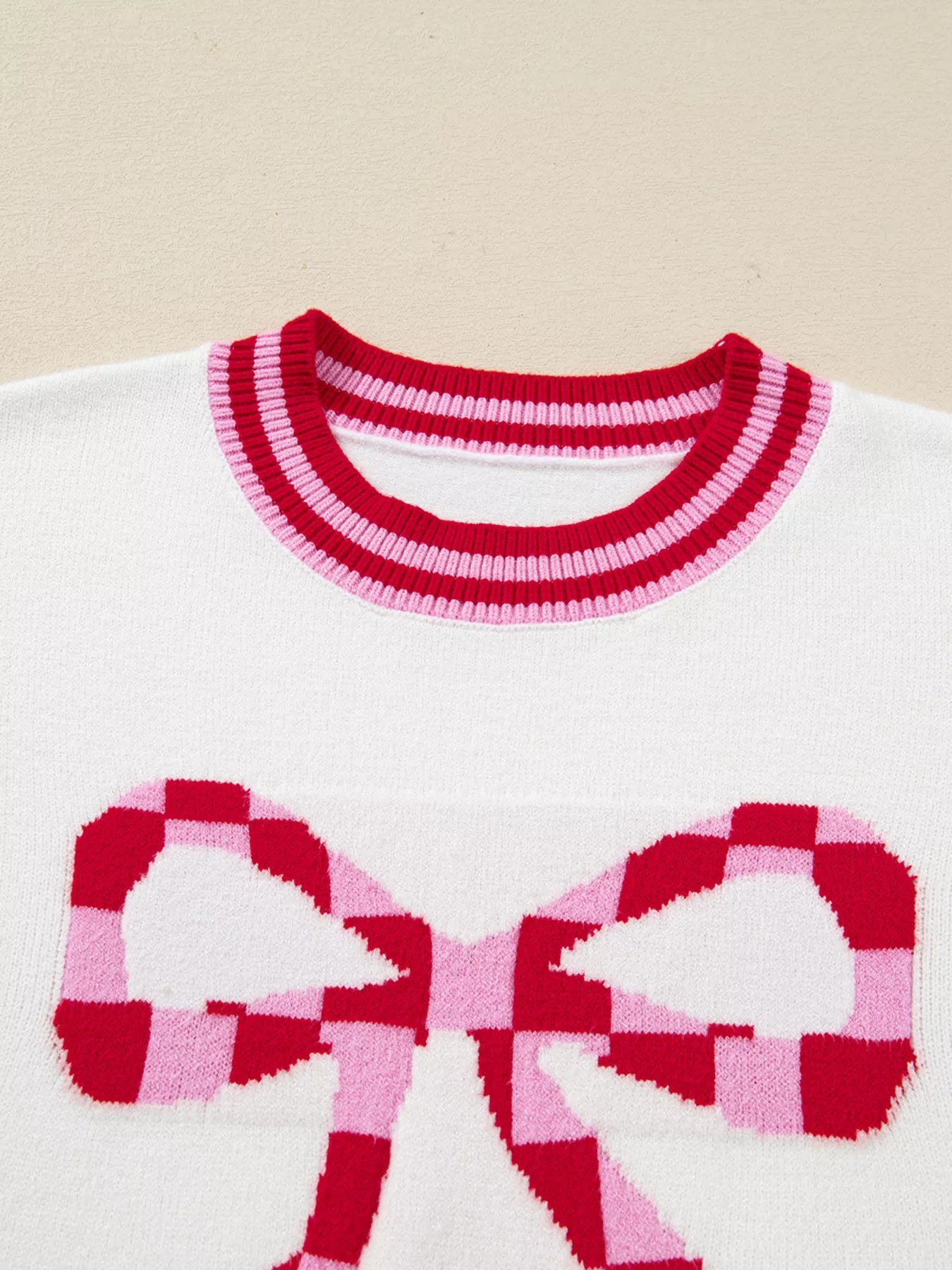 White sweater with red and pink checkered bow graphic and striped neckline.