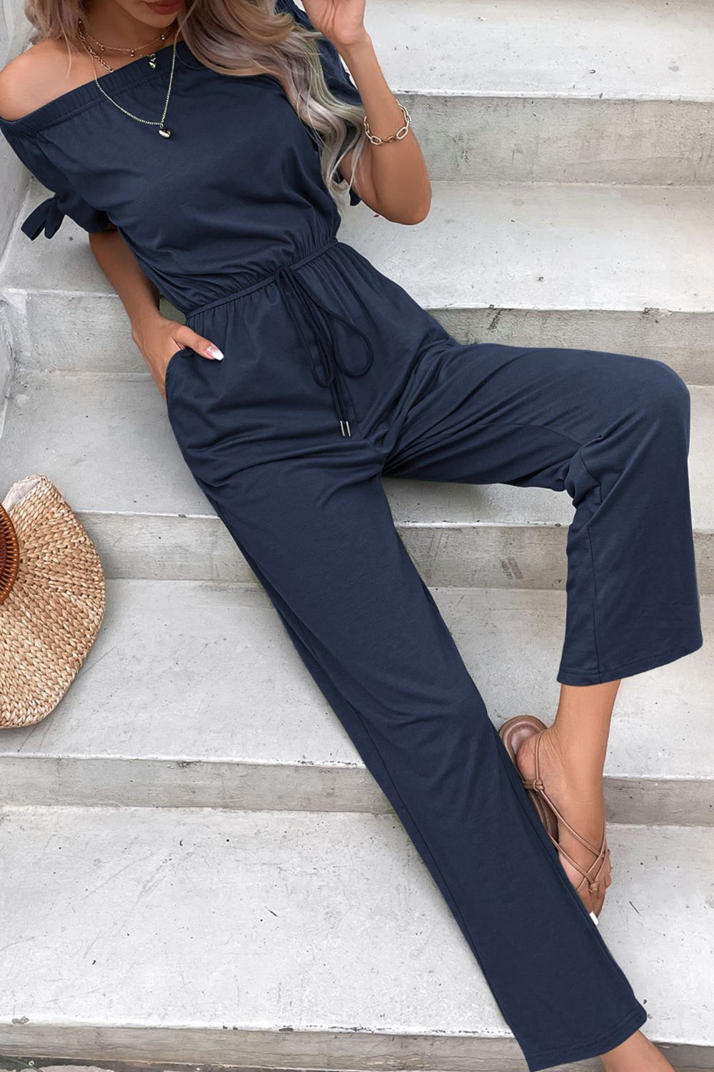 Off-the-shoulder navy jumpsuit with tie sleeves, drawstring waist, and pockets.