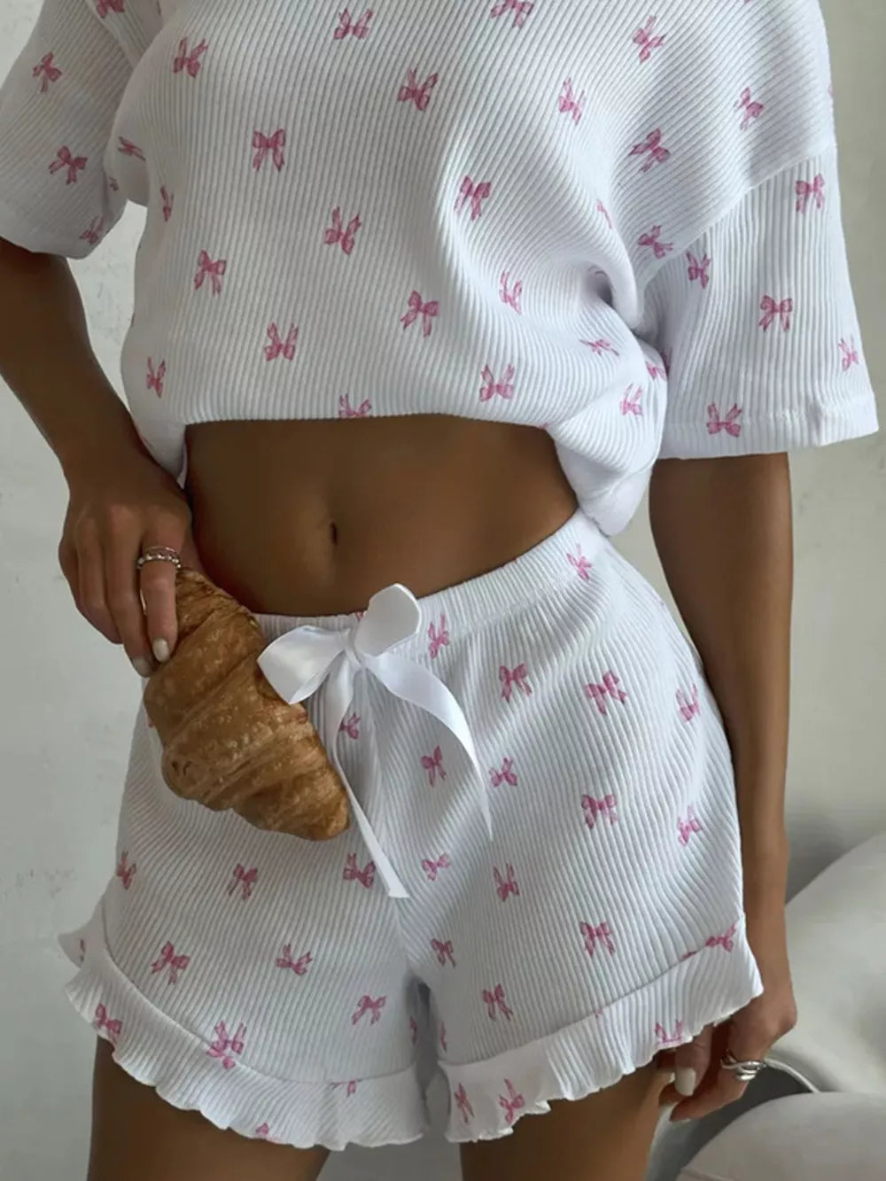 White knit lounge set with pink bow print, cropped top, and ruffled shorts with drawstring.
