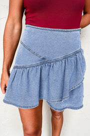 Asymmetrical denim ruffle mini skirt in medium wash cotton with tiered ruffles and a raw hem.