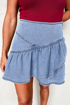Asymmetrical denim ruffle mini skirt in medium wash cotton with tiered ruffles and a raw hem.