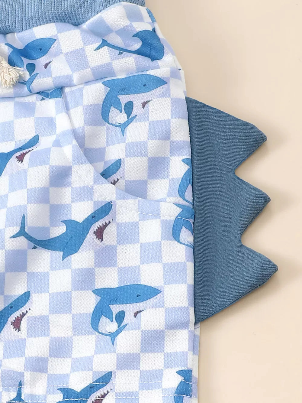 Baby blue checkered shorts with blue shark print and dinosaur tail detail.