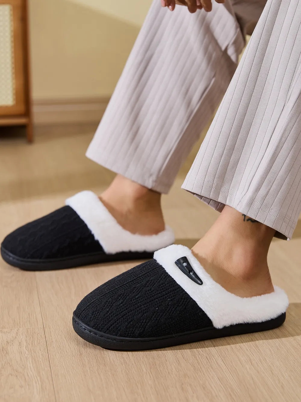 Black cable knit shearling house slippers with white faux fur trim and a decorative button detail.