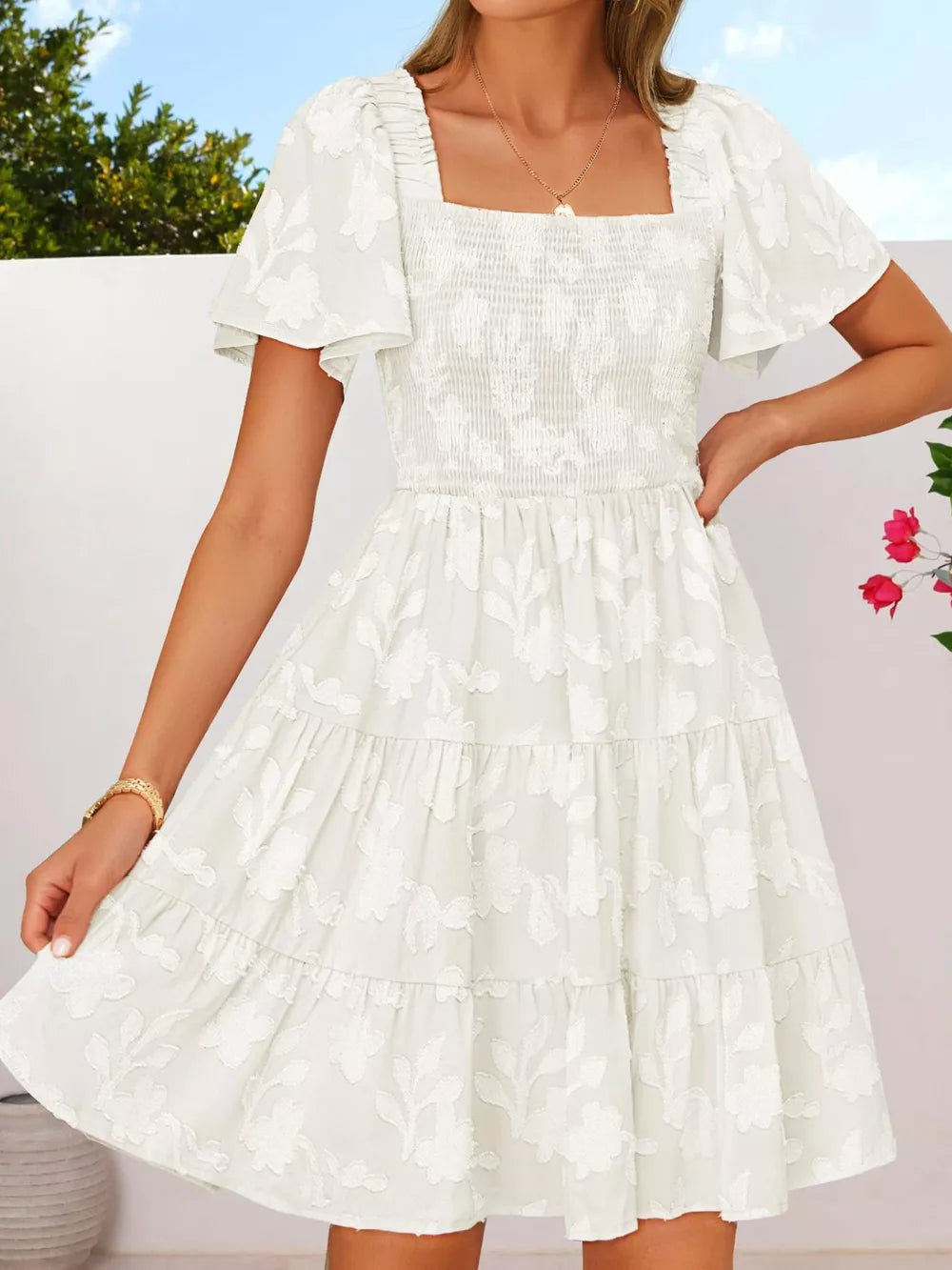 White smocked mini dress with tiered skirt, flutter sleeves, and floral embroidery.