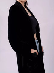 Black velvet longline trench coat with draped lapels and pockets.