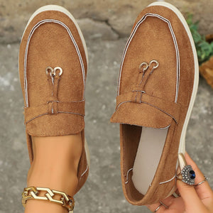 Brown flat slip-ons with decorative strap, white piping, and gold hardware.