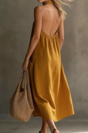 Mustard halter midi dress with pockets and open back, held by a model with a straw bag.